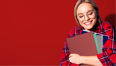 A woman with blonde hair and glasses smiles while holding four multi-colored notebooks to her chest and standing in front of a red background. She has her eyes closed and is wearing a red flannel with white and blue stripes.